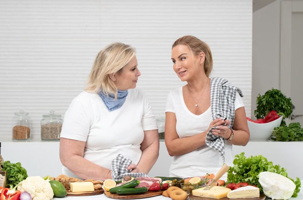 Two women in a kitchen preparing fresh vegetables and cheese with olive oil and spices.